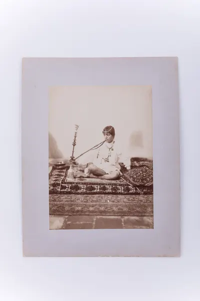 Fotografie de atelier ce prezintă o femeie așezată pe covor și care fumează dintr-o pipă cu apă. Imaginea este realizată pe hârtie albuminată, după un clișeu pe sticlă și este lipită pe un carton gros — تصویر رکورد یوروپیانا