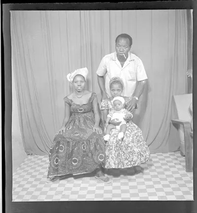 Four people, two women wearing dresses, head-wraps, and earrings sitting, one with a baby in a white outfit, and a man wearing a white shirt and a wrist watch with a cigarette in his mouth standing fa — تصویر رکورد یوروپیانا