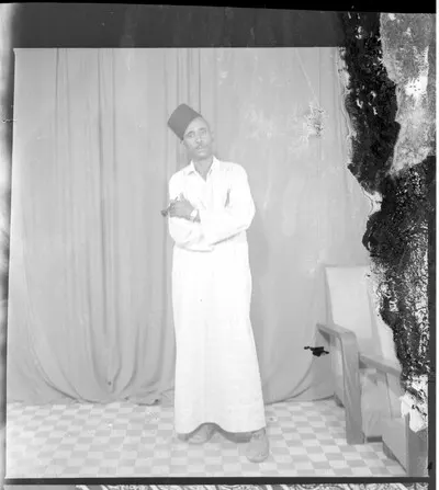 A man wearing a large white boubou, a black fez, and a wrist watch standing in profile in the studio. — تصویر رکورد یوروپیانا
