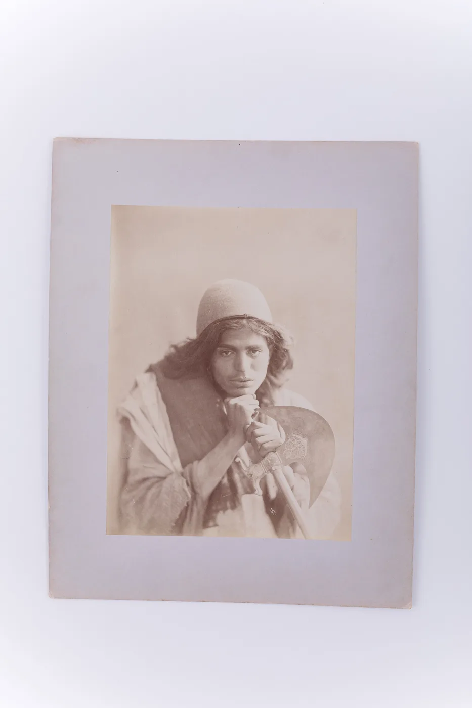 Fotografie de atelier ce prezintă un băiat dervish ce ține în mână o secure cu modele orientale. Imaginea este realizată pe hârtie albuminată, după un clișeu pe sticlă și este lipită pe un carton gros — تصویر رکورد یوروپیانا