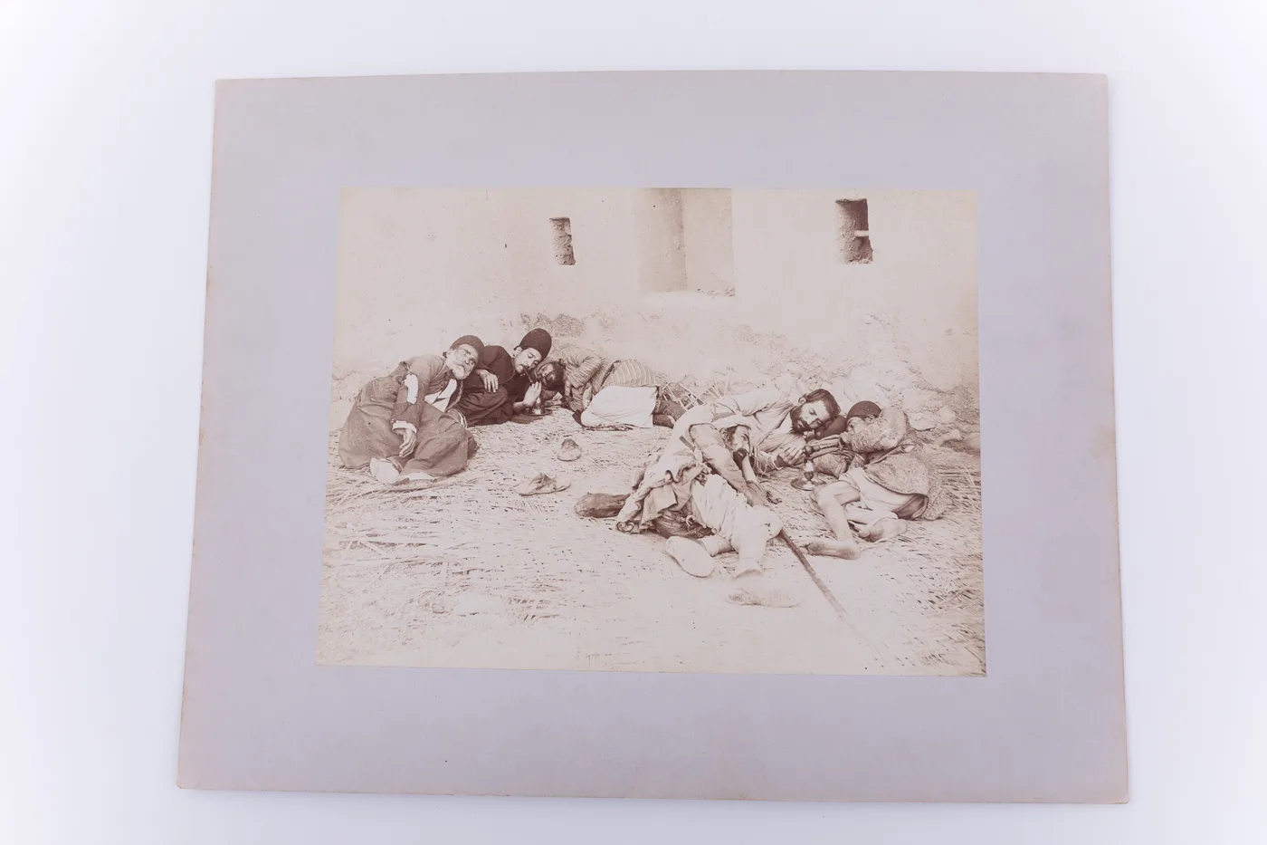 Fotografie de exterior ce prezintă un grup de bărbați care fumează opium. Acestia sunt surprinși întinși pe rogojini, în transă. Imaginea este realizată pe hârtie albuminată, după un clișeu pe sticlă  — تصویر رکورد یوروپیانا