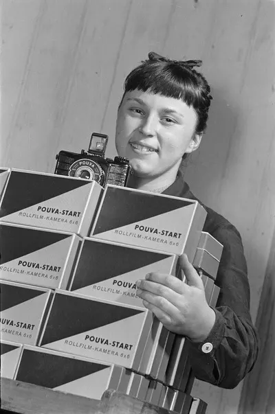 Freital. Karl Pouva KG [Camera Plant]. Employee with a stack of folding boxes for the rolling film camera “Pouva Start,” on which a camera unpacked — تصویر رکورد یوروپیانا