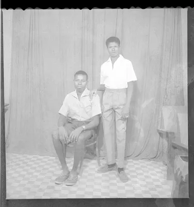 Two young people, one wearing a grey shirt, black shorts, and a wrist watch sitting and the other wearing a white short-sleeved shirt and grey pants standing, both face-forward in the studio. — تصویر رکورد یوروپیانا