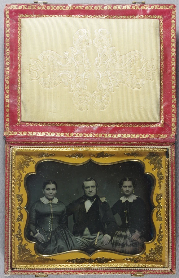 Bronze plate set in tortoise shell case designed to look like a book. Carved scene of riders in a forest on the front. Hand coloured daguerreotype of a seated group portrait of a man and two women. Wo — تصویر رکورد یوروپیانا