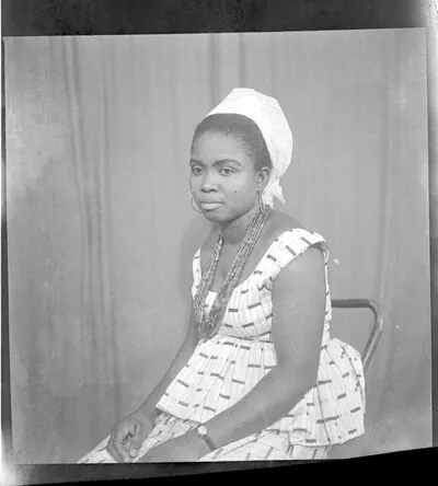 A woman wearing a dress, a white head-wrap, earrings, a necklace, and a wrist watch sitting in profile on a chair in the studio. — تصویر رکورد یوروپیانا