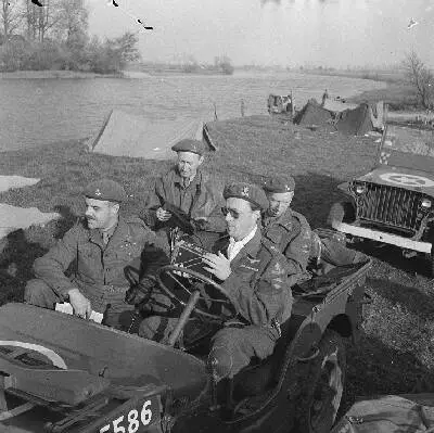 De Prins met camera in de hand bij een bivak aan de IJssel. Onder de voorruit van de tweede jeep is de standaard van Prins Bernhard zichtbaar. — تصویر رکورد یوروپیانا